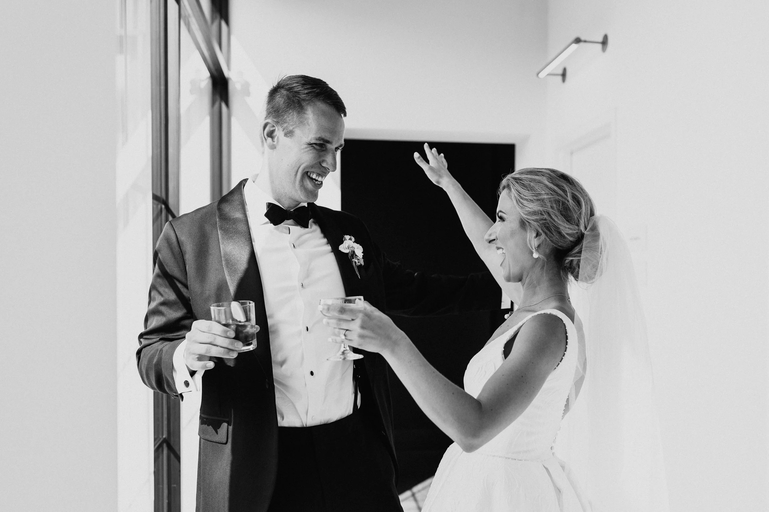 Black and white photo of a bride and groom celebrating with drinks, smiling and making a toast, indoor setting.