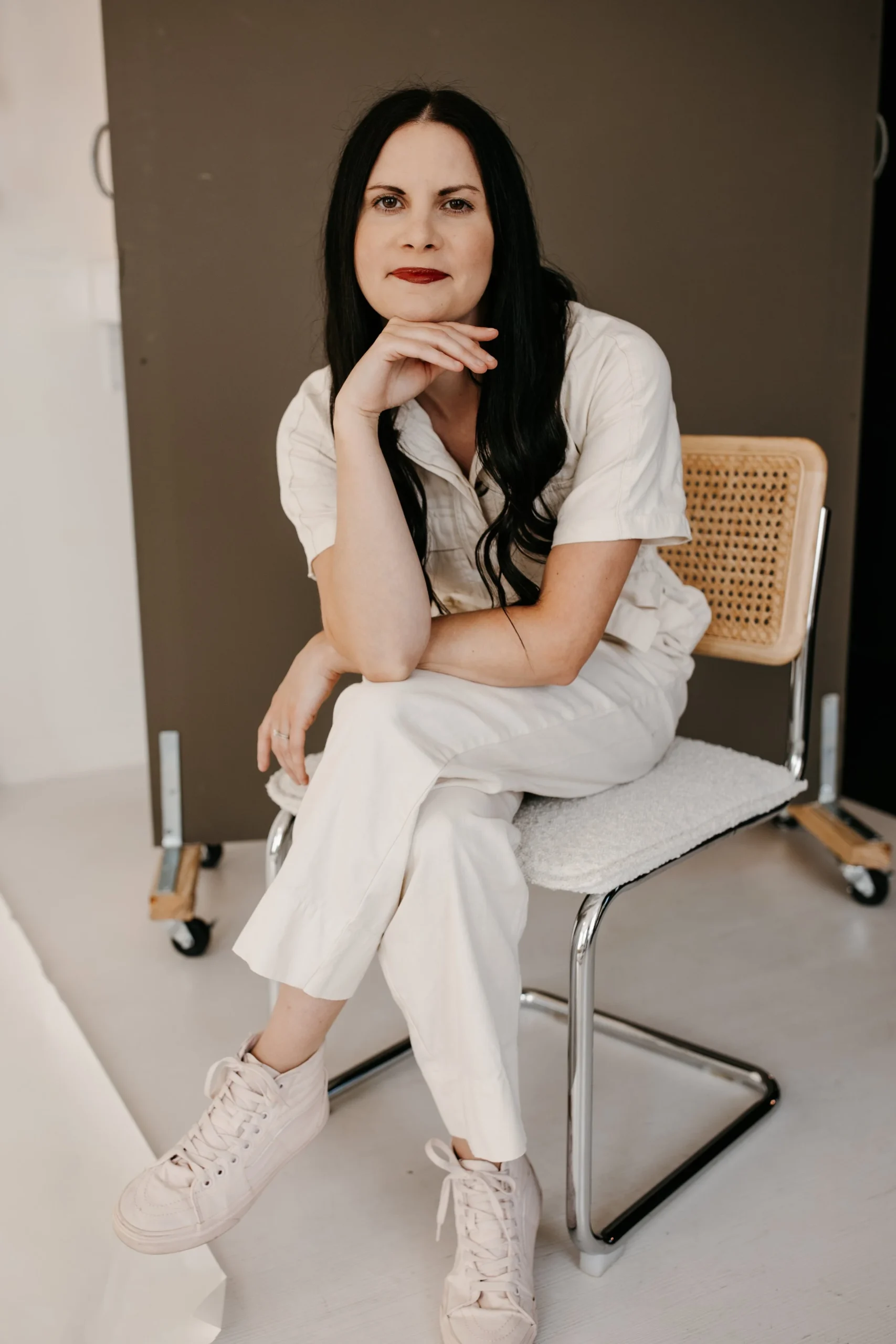 A woman with black hair and red lipstick sitting on a chair in a casual outfit, looking at the camera with her chin resting on her hand.