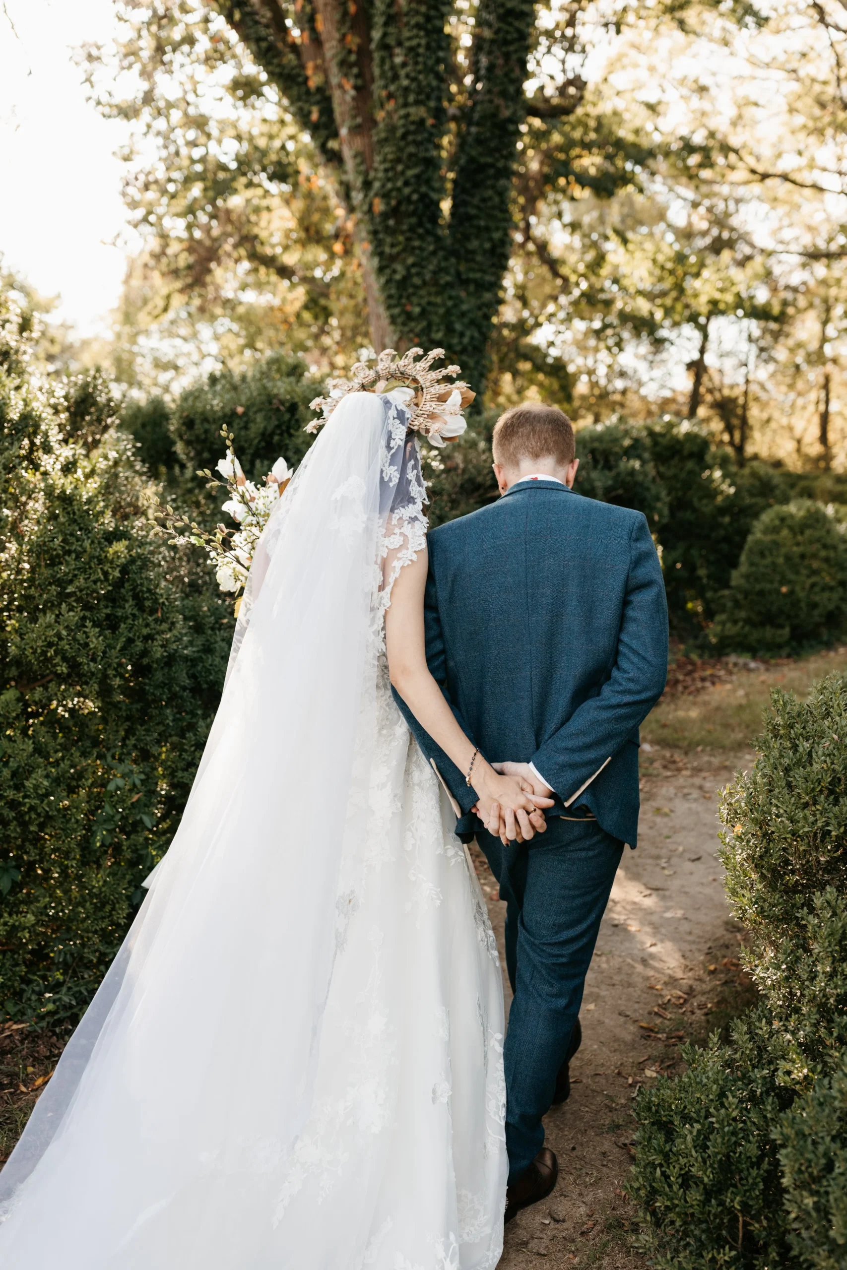 A bride and groom holding hands and walking into a garden during a wedding, with the bride wearing a long white lace gown and a veil, and a floral crown, while the groom is dressed in a blue suit.